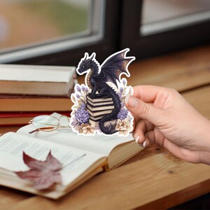 May include: A black dragon with gold accents sits on a stack of books. The dragon is surrounded by purple and white flowers. The sticker is on a book.