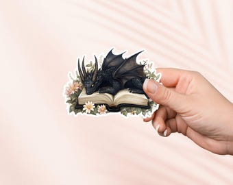 Puede incluir: Un dragón negro con alas enroscadas dormido sobre un libro abierto con detalles florales.