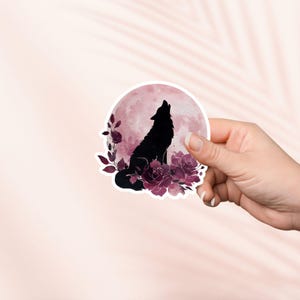 Peut inclure: Un autocollant rond représentant une silhouette noire de loup hurlant devant une lune rose. Le loup est entouré de roses et de feuilles rouge foncé. L'autocollant est tenu dans une main.