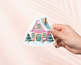 May include: A sticker featuring a pink and white bookstore with a snow-covered roof and two Christmas trees. The bookstore has a blue door with a heart and the word "Bookstore" above it.