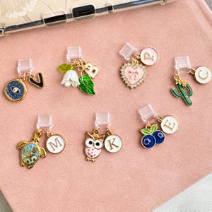 May include: A collection of seven gold-tone metal charms with clear plastic clips. The charms feature various designs including an evil eye, a white flower with a green leaf and letter B, a pink heart with letter P, a green cactus with letter C, a sea turtle with letter M, an owl with letter K, and blue berries with letter E.