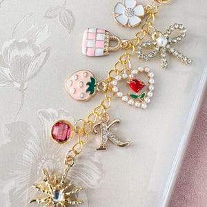 Op de afbeelding: Een goudkleurige sleutelhanger met meerdere decoratieve bedels. Bedels zijn onder meer een aardbei, een handtas, een bloem, een strik, een hart met een roos, de letter "K" en een zonnestraal. De bedels zijn in verschillende kleuren, waaronder roze, wit en rood.