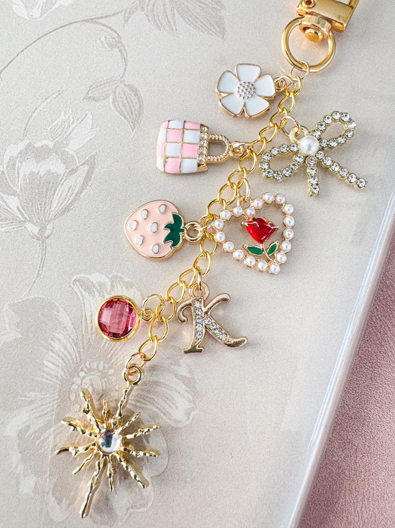 Op de afbeelding: Een goudkleurige sleutelhanger met meerdere decoratieve bedels. Bedels zijn onder meer een aardbei, een handtas, een bloem, een strik, een hart met een roos, de letter "K" en een zonnestraal. De bedels zijn in verschillende kleuren, waaronder roze, wit en rood.