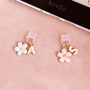 Puede incluir: Dos colgantes con forma de flor con detalles dorados y las letras "A" y "S". Los colgantes son blancos y rosas con un borde dorado. Los colgantes están unidos a un clip de plástico transparente. El fondo es una superficie rosa claro.