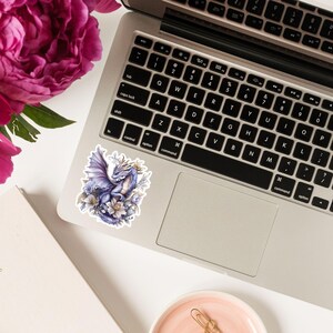 May include: A sticker featuring a purple dragon with gold accents, surrounded by white flowers. The dragon is depicted with wings spread and a gentle expression.