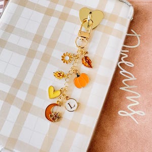 Peut inclure: Porte-clés doré avec un fermoir en forme de cœur et plusieurs breloques. Les breloques comprennent un tournesol, une feuille, une citrouille, un cœur, un croissant de lune et un cercle avec la lettre "L". Le porte-clés est sur un fond à carreaux.
