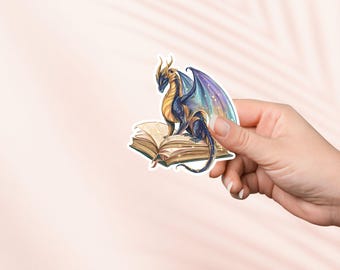 May include: A sticker featuring a fantasy dragon with blue, gold, and purple wings, perched atop an open book. The dragon has a detailed design, and the book has aged pages. The sticker is held against a soft pink background.