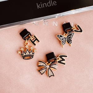 Op de afbeelding: Drie decoratieve bedels met zwarte en gouden accenten. Eén bedel heeft een kroon met heldere strass-steentjes, een andere een vlinder en de derde een strik met strass-steentjes. Elke bedel heeft een zwarte rechthoekige bovenkant en een goudkleurige letter.