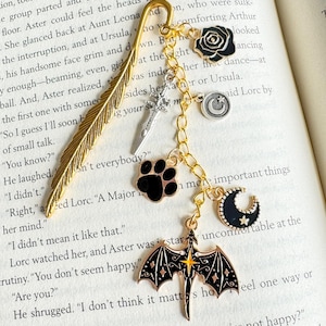 May include: A gold-colored metal bookmark with a feather design, adorned with charms. Charms include a black rose, a sword, a paw print, a crescent moon, a smiley face, and a dragon. The bookmark is on a book.