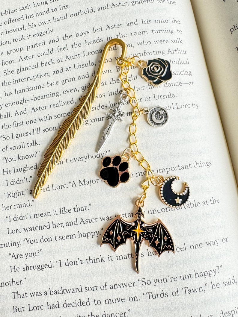 May include: A gold-colored metal bookmark with a feather design, adorned with charms. Charms include a black rose, a sword, a paw print, a crescent moon, a smiley face, and a dragon. The bookmark is on a book.