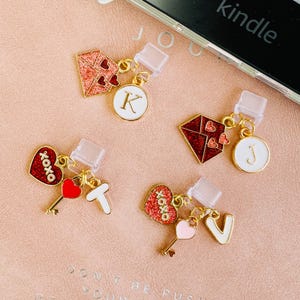 Peut inclure: Quatre breloques décoratives pour téléphone avec des accents dorés. Chaque breloque présente un design différent : un cœur rouge pailleté avec "xoxo", une forme de diamant rouge, un cercle blanc avec une lettre et une clé. Les breloques sont fixées à une prise de téléphone transparente.