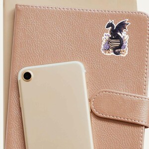 May include: A black dragon with purple accents sits on a stack of books with purple flowers and green leaves around it. The sticker is on a brown leather notebook.