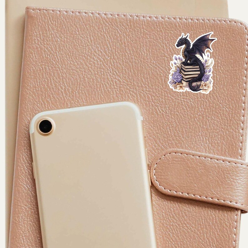 May include: A black dragon with purple accents sits on a stack of books with purple flowers and green leaves around it. The sticker is on a brown leather notebook.