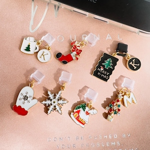 May include: A collection of Christmas-themed charms with gold-colored accents. The charms include a mug with a Christmas tree, a stocking, a mitten, a snowflake, a house, a Christmas tree, and letters. The background is a pink surface.