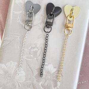 Op de afbeelding: Drie hartvormige bedels in zilver, zwart en goud, elk bevestigd aan een ketting en sluiting. De bedels worden getoond op een bloemmotief achtergrond, wat wijst op een decoratief of accessoire thema.