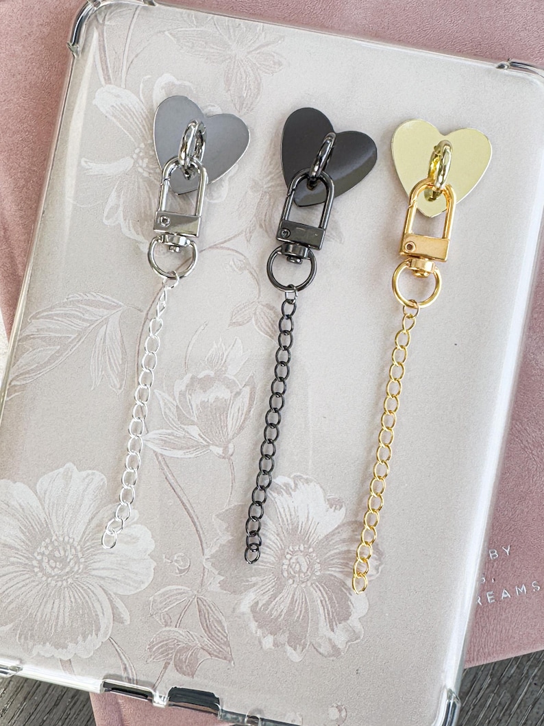 Op de afbeelding: Drie hartvormige bedels in zilver, zwart en goud, elk bevestigd aan een ketting en sluiting. De bedels worden getoond op een bloemmotief achtergrond, wat wijst op een decoratief of accessoire thema.