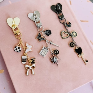 May include: Three decorative keychains with heart-shaped clasps. One is gold-toned with a purse, butterfly, coffee cup, and bow charms. The second is silver-toned with a butterfly, rose, and book charms. The third is black with heart, sun, and key charms.