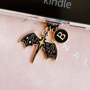 Może przedstawiać: Złoty i czarny wisiorek w kształcie smoka z żółtą gwiazdą, przymocowany do czarnego sznurka, oraz czarny okrągły wisiorek z literą "B". Ozdoby znajdują się na różowej powierzchni.