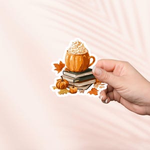 May include: A sticker featuring a pumpkin-shaped mug filled with whipped cream, resting on a stack of books. Small pumpkins and autumn leaves surround the mug and books, creating a fall theme.
