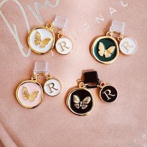 May include: Four decorative charms with gold-coloured accents. Each charm features a butterfly design and the letter "R". The charms are in white, green, pink, and black, with a gold-coloured butterfly in the centre.
