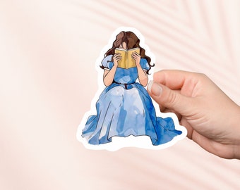 Puede incluir: Una pegatina con una mujer en un vestido azul sentada y leyendo un libro. La mujer tiene el pelo castaño y lleva un vestido azul con un cinturón blanco. El libro es amarillo y tiene una portada blanca.