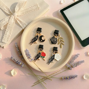 May include: A selection of black and gold earrings featuring raven, crescent moon, rose, and leaf designs. The earrings are presented on a cream plate, with lavender sprigs and a gift box visible in the background.