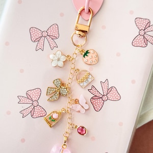 Könnte beinhalten: Ein goldfarbenes Charm-Armband mit verschiedenen Anhängern, darunter eine Erdbeere, eine Blume, eine Schleife, ein Schmetterling und ein Herz. Die Anhänger sind an einer Kette und einem herzförmigen rosa Verschluss befestigt. Der Hintergrund ist ein rosa Notizbuch mit Schleifenmustern.