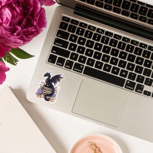 May include: A black dragon with purple wings sits on top of a stack of books. The dragon is surrounded by purple and white flowers. The sticker is on a laptop.