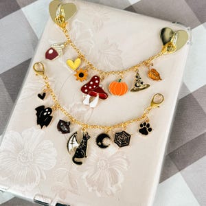 May include: Two gold-tone metal charm bracelets featuring seasonal motifs. The top bracelet includes an acorn, heart, sunflower, mushroom, pumpkin, witch hat, and leaf. The bottom bracelet features a ghost, heart, cat with wings, moon, spiderweb, and paw print.