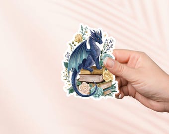 Puede incluir: Una pegatina con un dragón azul con alas turquesas posado sobre una pila de libros. El dragón está rodeado de rosas amarillas y turquesas y vegetación. La pegatina está sujeta por una mano sobre un fondo rosa claro.