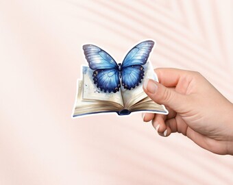 Puede incluir: Una mariposa azul con marcas negras está posada sobre un libro abierto con páginas envejecidas. El libro está sostenido en una mano.
