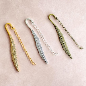 May include: Four metal feather bookmarks in gold, silver, and bronze tones. Each bookmark features a feather design and a chain. The bookmarks are arranged diagonally on a pink surface.