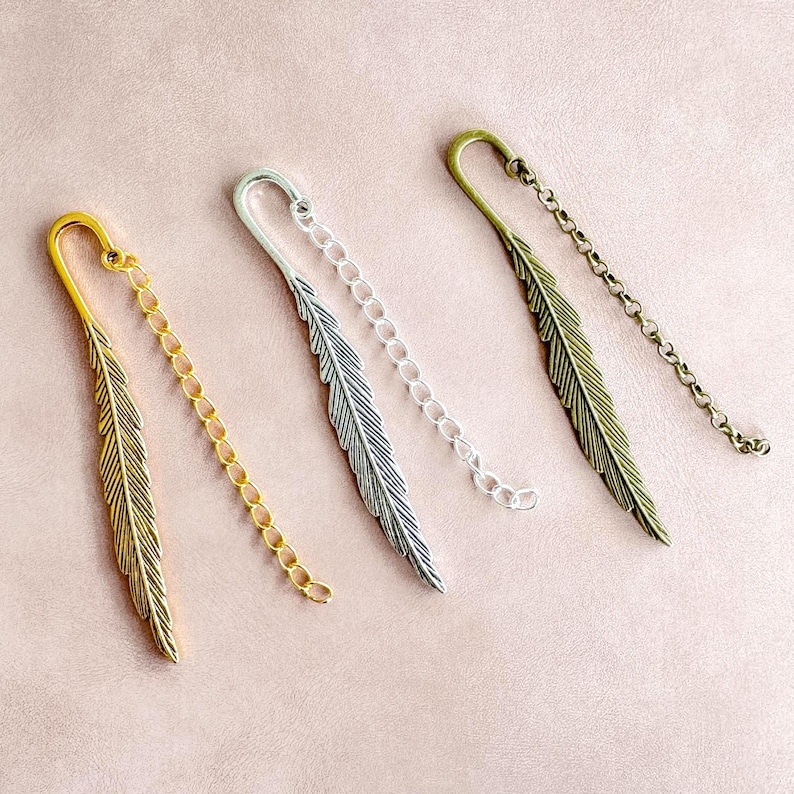 May include: Four metal feather bookmarks in gold, silver, and bronze tones. Each bookmark features a feather design and a chain. The bookmarks are arranged diagonally on a pink surface.