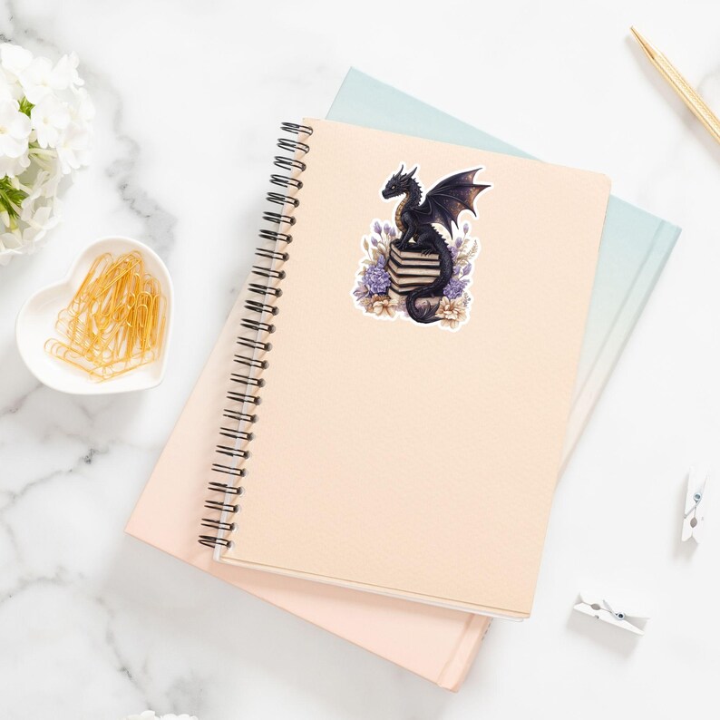 May include: A black dragon with purple flowers and a stack of books. The dragon is sitting on top of the books. The image is a sticker.