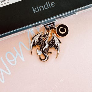 May include: A dragon charm in black and white with gold detailing, suspended from a Kindle. The dragon features intricate wing patterns and a curled tail. A circular charm with a black 'C' is also present. The background is pale pink.