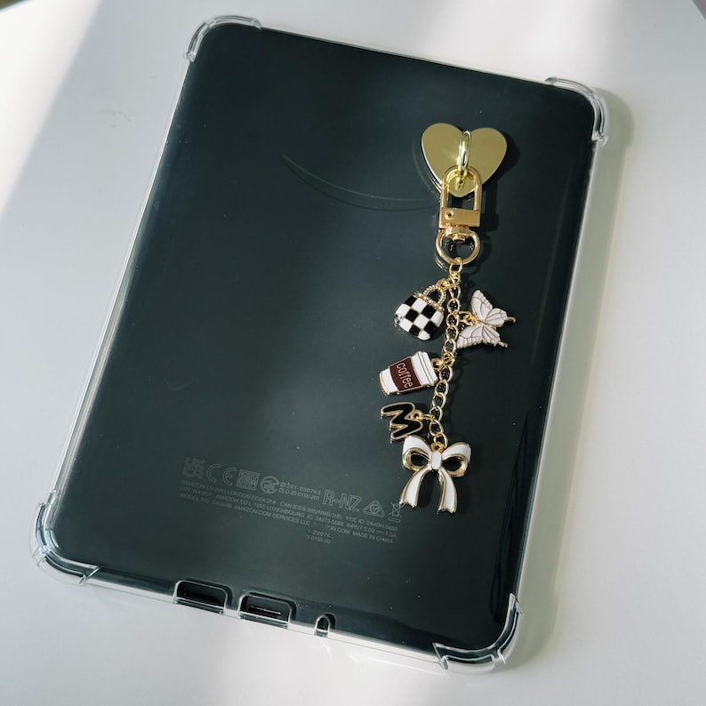 Op de afbeelding: Zwarte tablet in een doorzichtige hoes. Een goudkleurige hartvormige bedel hangt aan de bovenkant, met een ketting van bedels: een geruite tas, een vlinder, een koffiekopje en een strik. De tablet is ongeveer 20 cm lang.