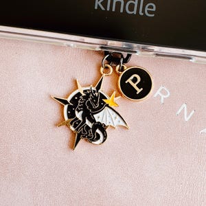 May include: A black and white dragon charm with gold accents and a gold starburst design. The charm is attached to a phone case with a black ring and a gold charm with the letter "P".