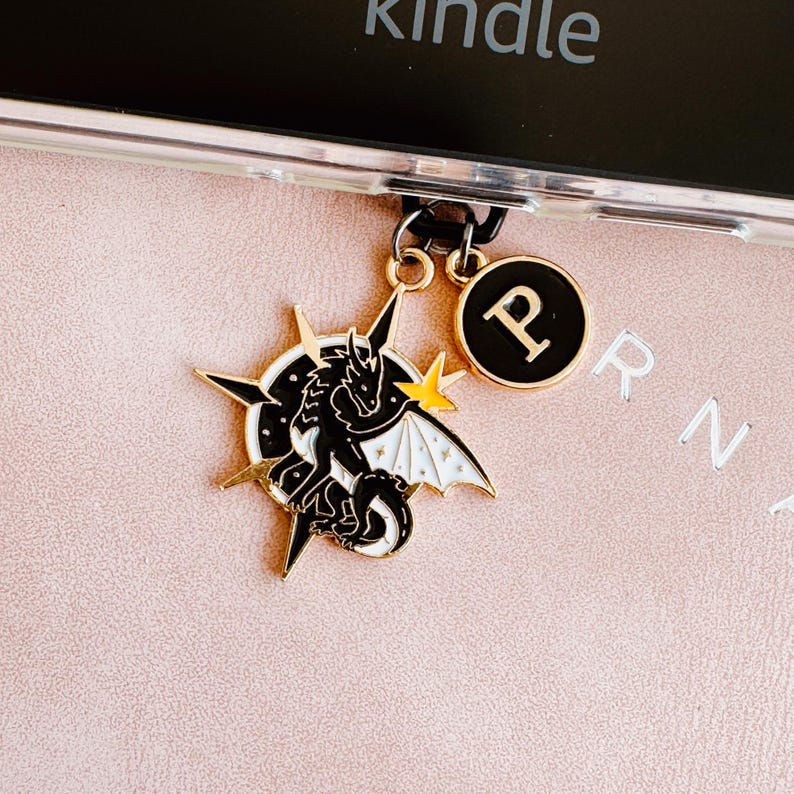 May include: A black and white dragon charm with gold accents and a gold starburst design. The charm is attached to a phone case with a black ring and a gold charm with the letter "P".