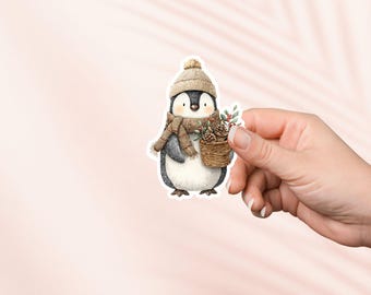 May include: A sticker featuring a watercolor illustration of a penguin wearing a knit hat, scarf, and carrying a basket of pinecones and berries. The penguin is primarily black and white with brown accessories. The sticker is held by a hand against a pink background.