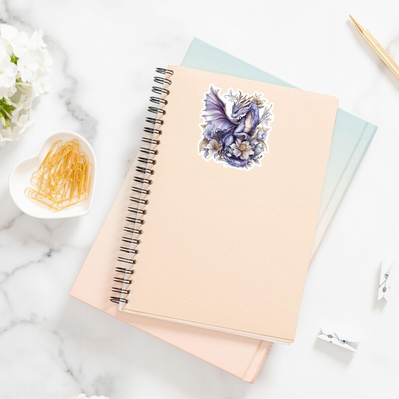 May include: A sticker featuring a purple dragon with wings spread, surrounded by white flowers. The dragon has a crown on its head and is set against a white background.