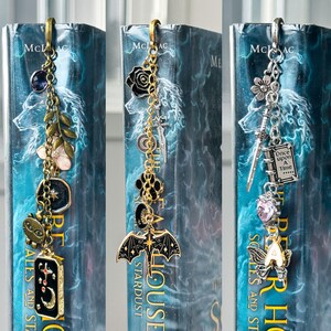 May include: Three decorative metal bookmarks with dangling charms. One has a purple gem, a flower, and a star. Another features a black rose, a bat, and a crescent moon. The third includes a flower, a sword, and a charm that says "Once Upon A Time".
