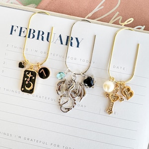 May include: Three decorative bookmarks with charms. One gold bookmark has a black and gold charm with a crescent moon and stars, and a black charm with a gold symbol. Another silver bookmark has a dragon charm and a black heart charm. The third gold bookmark has a cross charm.