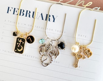 May include: Three decorative bookmarks with charms. One gold bookmark has a black and gold charm with a crescent moon and stars, and a black charm with a gold symbol. Another silver bookmark has a dragon charm and a black heart charm. The third gold bookmark has a cross charm.