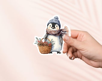May include: A watercolor sticker of a penguin wearing a blue knit hat and scarf, carrying a woven basket filled with pinecones and greenery. The penguin has a black and white body and is smiling. The sticker is held by a hand against a pink background.