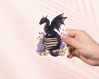Op de afbeelding: Een zwarte draak met gouden accenten zit op een stapel boeken omringd door paarse en crèmekleurige bloemen.