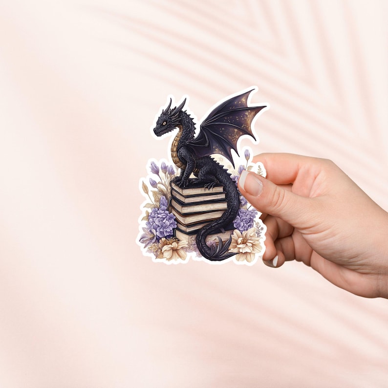 May include: A black dragon with gold accents sits on a stack of books surrounded by purple and cream flowers.