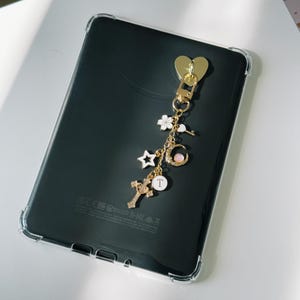 Op de afbeelding: Een zwarte tablet met een doorzichtige beschermhoes, met een goudkleurige hartvormige bedel en een decoratieve ketting met diverse bedels, waaronder een kruis, een ster en een maan. De bedels zijn goudkleurig met witte en roze accenten.