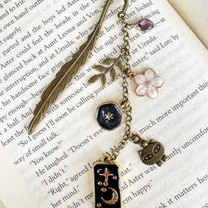May include: A decorative bookmark with a bronze feather design and dangling charms. Charms include a purple gem, a white flower, a black pendant with a star, and a cat charm. The bookmark is on a book.
