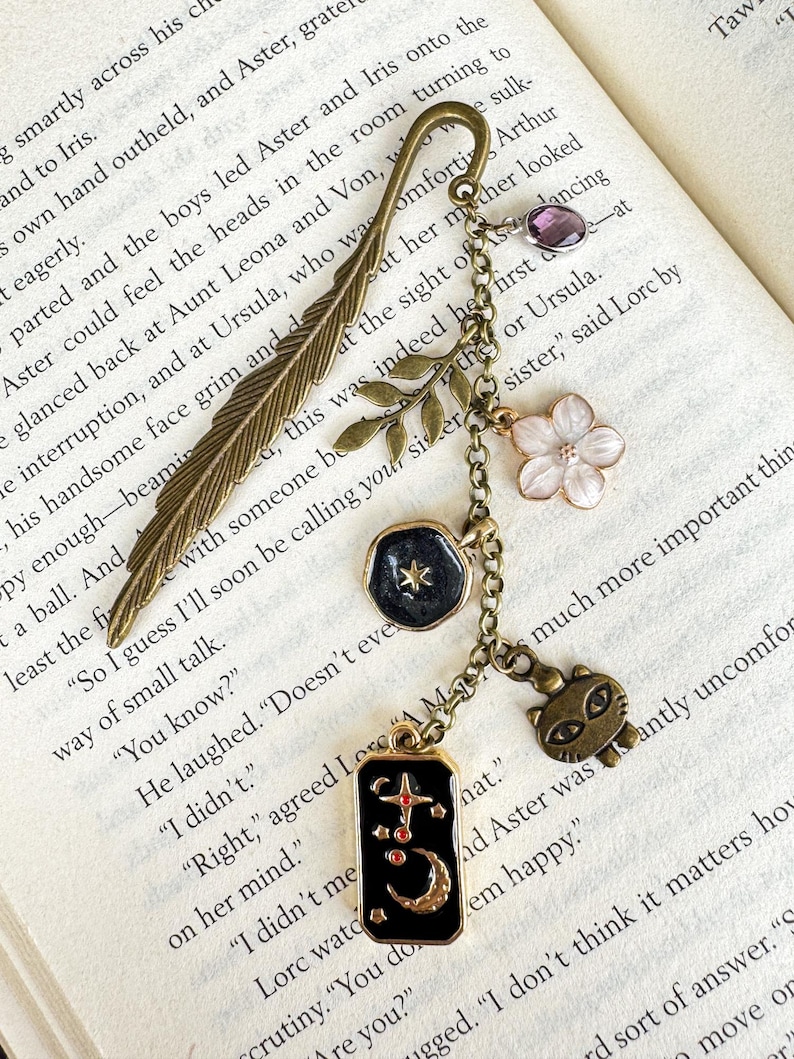 May include: A decorative bookmark with a bronze feather design and dangling charms. Charms include a purple gem, a white flower, a black pendant with a star, and a cat charm. The bookmark is on a book.