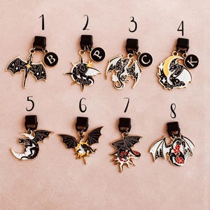 May include: Collection of eight dragon-themed charms with black clasps. Each charm features a different dragon design in black, white, and gold, with some including red accents. Each charm has a letter on a small circular charm.